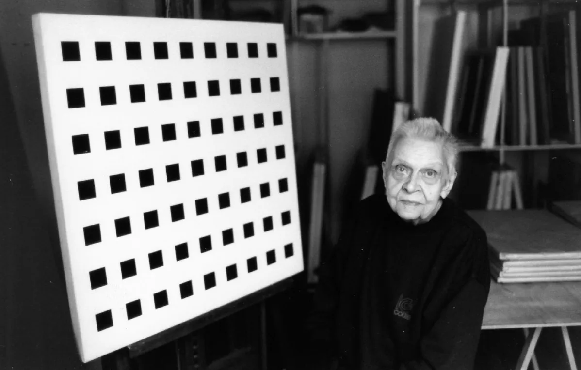Retrato de Aurélie Nemours en su taller: mujer mayor de pelo blanco corto y jersey oscuro, sentada junto a un lienzo blanco con una retícula de cuadrados negros.