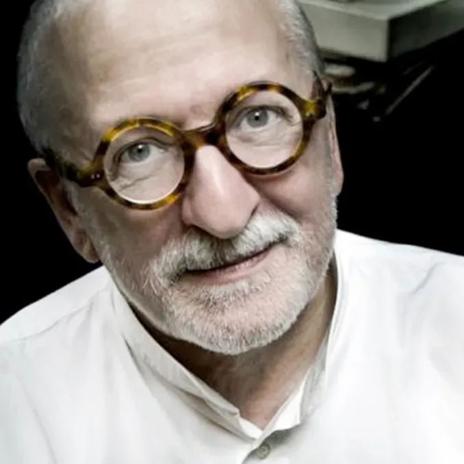 Retrato de Ennio Chiggio: hombre mayor de barba blanca y gafas redondas de carey, con camisa blanca sobre fondo oscuro.
