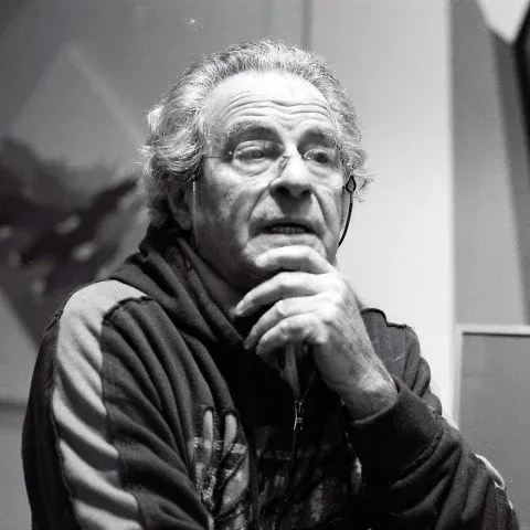 Retrato de Franco Giuli: hombre mayor de pelo canoso y gafas, con la mano en el mentón y chaqueta deportiva oscura.