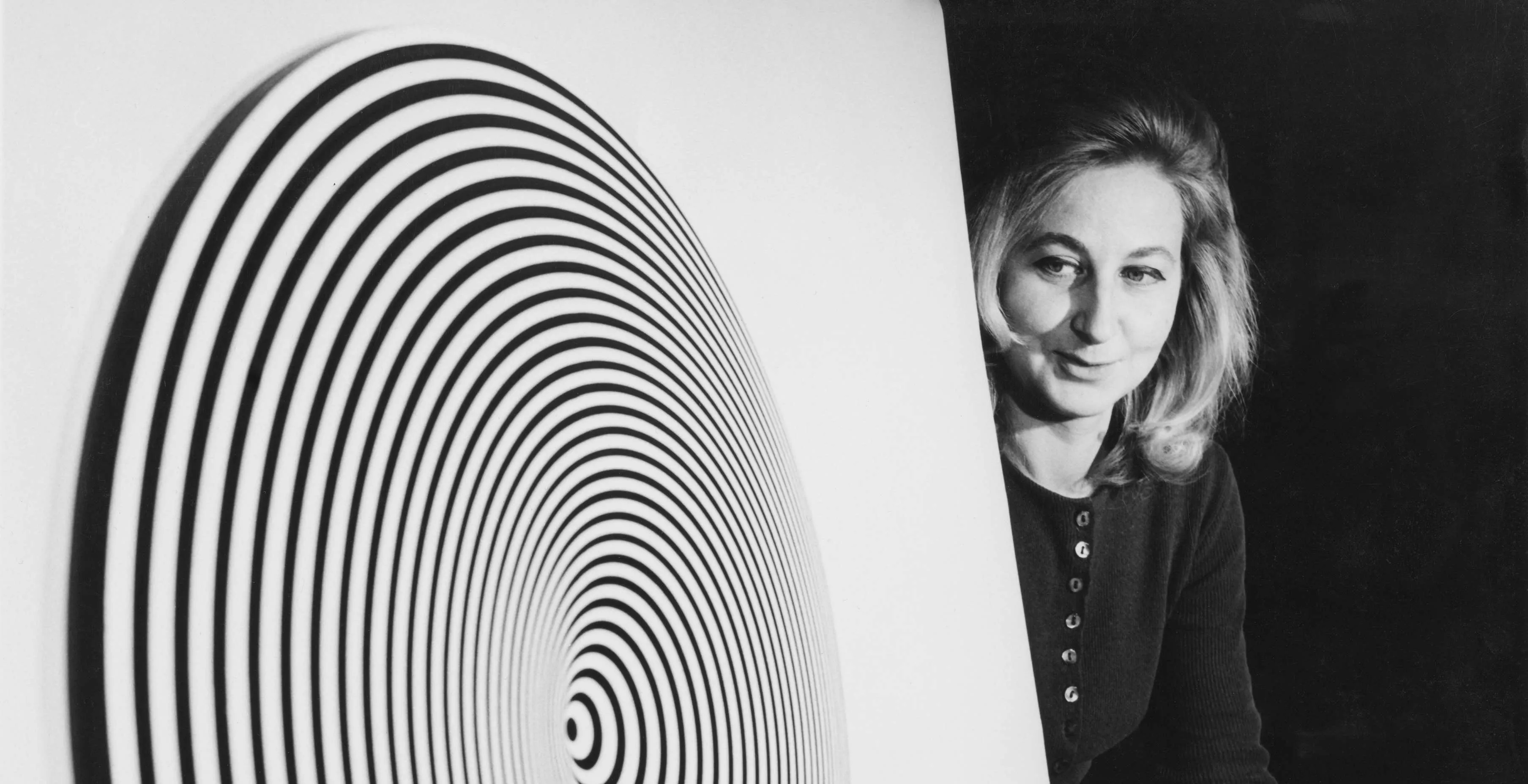 Retrato de Marina Apollonio: mujer joven de melena rubia y cárdigan oscuro, asomada tras un disco de círculos concéntricos blancos y negros.